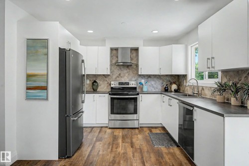Modern kitchen featuring white cabinetry, dark countertops, wood-finish flooring, stainless steel appliances, and a tiled backsplash - 3718 103B Street Nw, Edmonton, AB - Indoor Photo Showing Kitchen With Double Sink