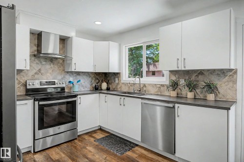Modern kitchen featuring white cabinetry, dark countertops, stainless steel appliances, a tile backsplash, and wood-finish flooring - 3718 103B Street Nw, Edmonton, AB - Indoor Photo Showing Kitchen With Double Sink