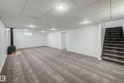 Spacious lower-level room featuring patterned carpet flooring, a black wood-burning stove on a tiled hearth, recessed lighting, and a drop ceiling - 3718 103B Street Nw, Edmonton, AB - Indoor Photo Showing Other Room