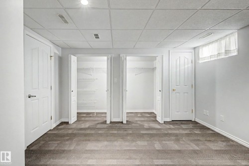 Carpeted room featuring two white paneled doors, two open closets with wire shelving, a window with curtain, and a dropped ceiling with recessed lighting - 3718 103B Street Nw, Edmonton, AB - Indoor Photo Showing Other Room