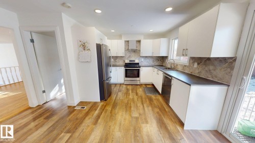 Modern kitchen featuring white cabinetry, stainless steel appliances, dark countertops, and wood-finish flooring - 3718 103B Street, Edmonton, AB 