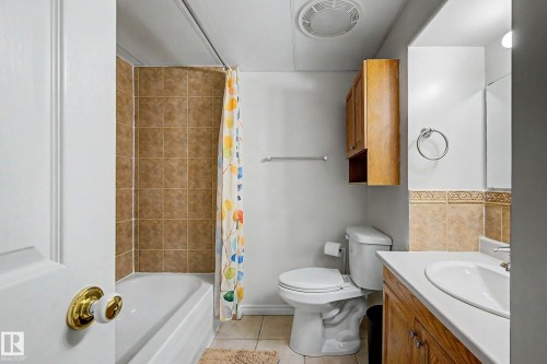 Bathroom featuring a tub/shower combination with brown square tile surround, a single vanity with an integrated sink and wood-finish cabinetry, a toilet, and a wall-mounted wood-finish cabinet - 3718 103B Street Nw, Edmonton, AB - Indoor Photo Showing Bathroom