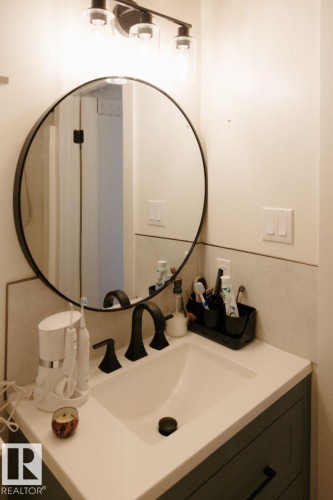 Bathroom vanity featuring a quartz-finish countertop with an integrated sink, matte black faucet, and a large round mirror - 10503A 109 Street, Westlock, AB - Indoor Photo Showing Bathroom