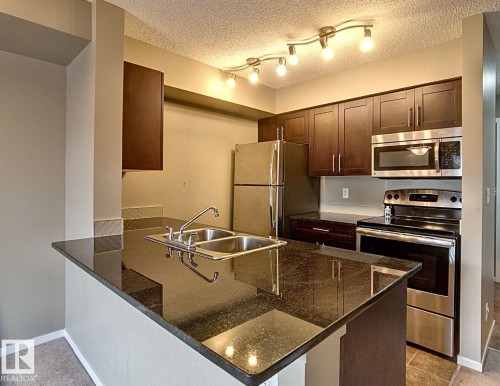 Kitchen featuring dark wood-finish cabinetry, stainless steel appliances, a double basin sink, track lighting, and dark stone-finish countertops - 328 3315 James Mowatt Trail, Edmonton, AB 