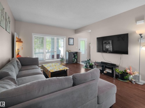 Spacious living area featuring warm wood-finish flooring, a large picture window with blinds, and neutral wall tones - 7827 22 Avenue, Edmonton, AB 