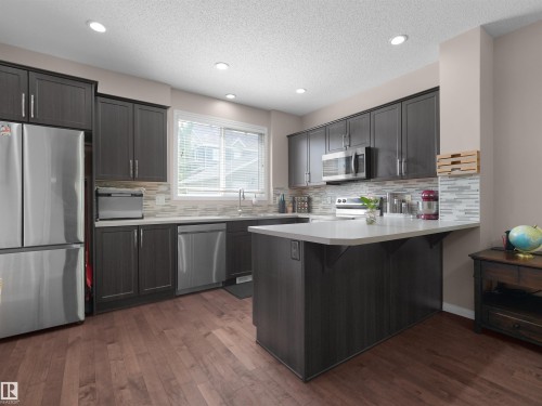 Modern kitchen featuring dark wood-finish cabinetry, stainless steel appliances, light-toned countertops, a multi-toned tile backsplash, and recessed lighting - 7827 22 Avenue, Edmonton, AB 
