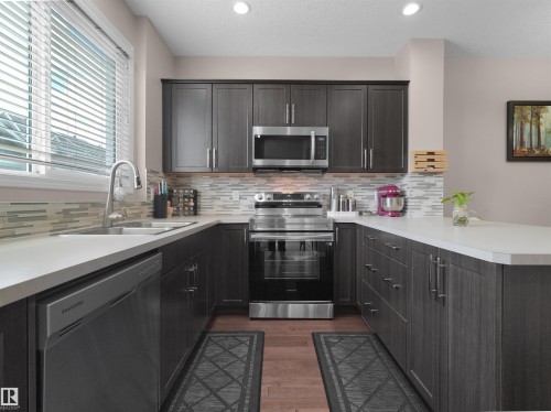 Modern kitchen featuring dark wood-finish cabinetry, stainless steel appliances, light countertops, and a mosaic tile backsplash - 7827 22 Avenue, Edmonton, AB 