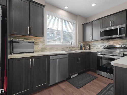 Contemporary kitchen featuring dark wood-finish cabinetry, stainless steel appliances, light countertops, a patterned tile backsplash, and wood-finish flooring - 7827 22 Avenue, Edmonton, AB 