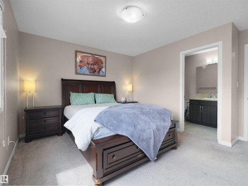 Carpeted bedroom featuring a dark wood-finish bed frame, two matching dark wood-finish nightstands, a ceiling-mounted light fixture, and an entry to a bathroom with a dark wood-finish vanity - 7827 22 Avenue, Edmonton, AB 
