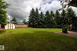 Expansive grassy yard featuring a wood privacy fence, mature evergreen trees, a detached storage shed, and a brick paver patio with exterior wood stairs - 