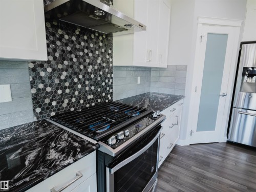 Modern kitchen featuring a stainless steel range with gas burners, a hexagonal mosaic tile backsplash, dark patterned countertops, white cabinetry, and wood-finish flooring - 2526 Wonnacott Loop, Edmonton, AB 