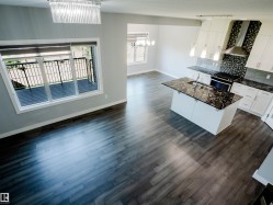 Expansive open-concept living area featuring wood-finish flooring, a contemporary kitchen island with a dark stone countertop, white cabinetry, and a mosaic tile backsplash - 
