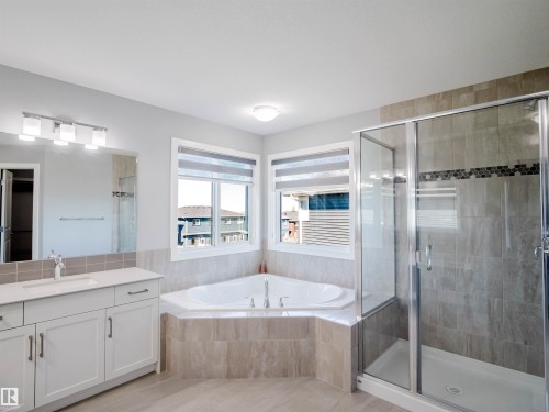 Spacious bathroom featuring a corner soaking tub, a separate glass-enclosed shower with tile surround, a single vanity with white cabinetry and a light-toned countertop, and dual windows with shades - 2526 Wonnacott Loop, Edmonton, AB 
