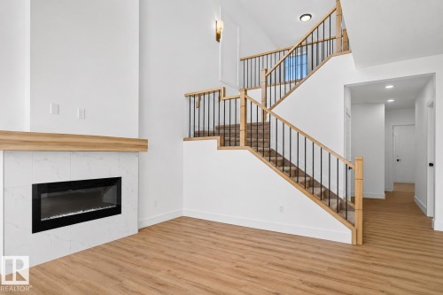 Open-concept living space featuring wood-finish flooring, a contemporary fireplace with white marble-look surround and wood mantle, and a staircase with natural wood handrails and black balusters - 63 Silverstone Drive, Stony Plain, AB 