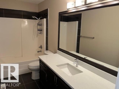 Contemporary bathroom featuring a dark wood-finish vanity with a white countertop and integrated sink, a large framed mirror, and a full bathtub with tiled surround - 805 Hodgins Road, Edmonton, AB 