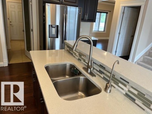 Kitchen island featuring a double basin stainless steel sink with a high-arch faucet, light-toned countertop, and a tiled backsplash - 805 Hodgins Road, Edmonton, AB 