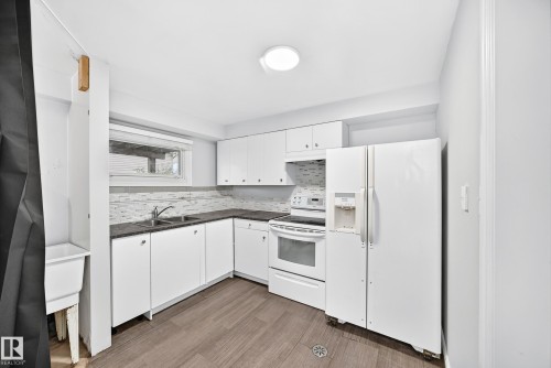 Kitchen featuring white cabinetry, dark countertops, a mosaic tile backsplash, wood-finish flooring, and a white appliance suite - 1103 56 Street, Edmonton, AB 