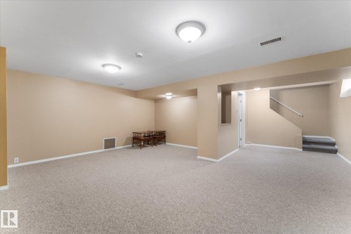 Spacious lower-level room featuring light neutral wall paint, light-toned carpet flooring, recessed lighting, and a partial staircase with a metal handrail - 15112 76A Avenue, Edmonton, AB 