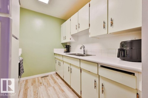 Galley kitchen featuring white cabinetry, light countertops, a stainless steel double sink, white tile backsplash, and wood-finish flooring - 17009 109 Street, Edmonton, AB 