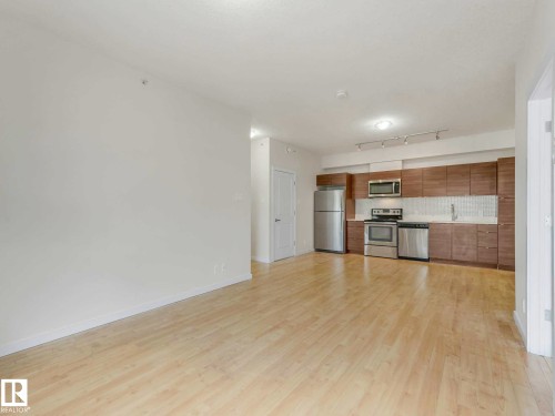 Open-concept living space featuring wood-finish flooring, a modern kitchen with wood-finish cabinetry, stainless steel appliances, and track lighting - 406 9519 160 Avenue, Edmonton, AB 