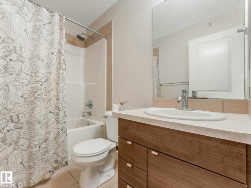 Bathroom featuring a wood-finish vanity with an integrated sink and chrome faucet, a large wall-mounted mirror, and a white bathtub with a shower and tile surround - 406 9519 160 Avenue, Edmonton, AB 