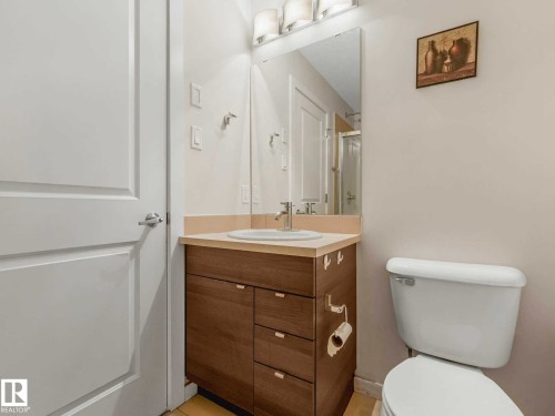 Bathroom featuring a wood-finish vanity with a light-toned countertop, an integrated sink, a wall-mounted mirror, and a modern light fixture - 406 9519 160 Avenue, Edmonton, AB 