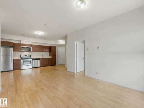 Open-concept living space featuring wood-finish flooring and a kitchen area with stainless steel appliances, dark wood-grain cabinetry, and a light-toned backsplash - 406 9519 160 Avenue, Edmonton, AB 
