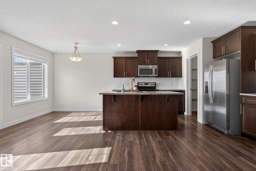 Open-concept kitchen and dining area featuring rich wood-finish flooring, a central island with a granite-style countertop, stainless steel appliances, dark wood cabinetry, and a subway tile backsplash - 5306 67 Street E, Beaumont, AB 