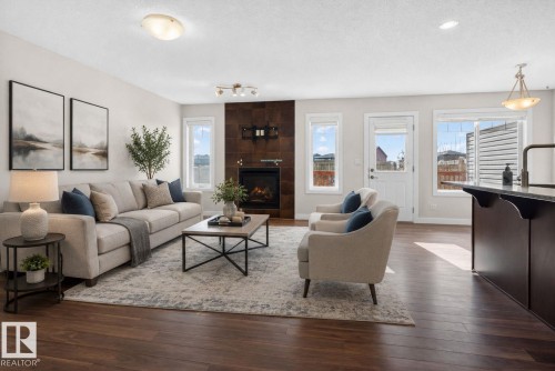 Open-concept living area featuring rich wood-finish flooring, a contemporary fireplace with a dark tile surround, and multiple windows - 5306 67 Street E, Beaumont, AB 