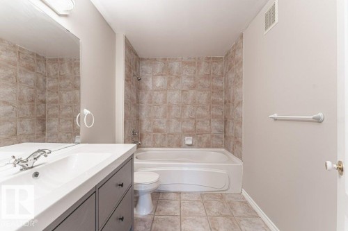 Bathroom featuring a built-in bathtub with tiled surround, a vanity with an integrated sink and storage drawers, a wall-mounted mirror, and tile flooring - 101 10421 42 Avenue, Edmonton, AB 