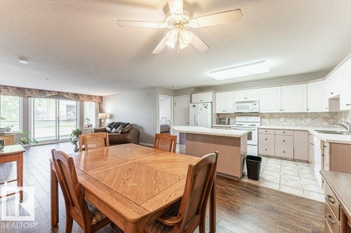 Open-concept living space featuring wood-finish flooring, a kitchen with white cabinetry and tiled backsplash, a kitchen island with a breakfast bar, and a dining area with a ceiling fan - 101 10421 42 Avenue, Edmonton, AB 
