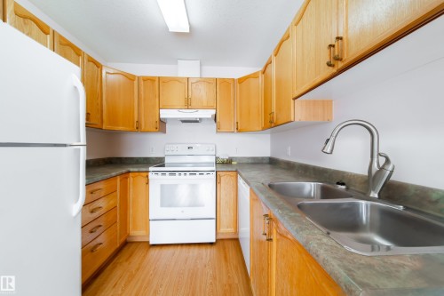 U-shaped kitchen featuring wood-finish cabinetry, a double basin stainless steel sink with a high-arc faucet, and wood-finish flooring - 120 13320 124 Street Nw, Edmonton, AB 