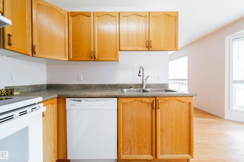 Kitchen featuring wood-finish cabinetry, a double basin sink with a gooseneck faucet, a white built-in dishwasher, and a wood-finish flooring - 120 13320 124 Street Nw, Edmonton, AB 