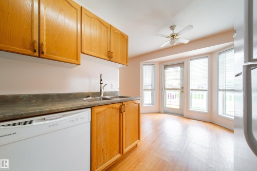 Kitchen area featuring wood-finish flooring, ample wood cabinetry, a dual basin stainless steel sink, and a white built-in dishwasher - 120 13320 124 Street Nw, Edmonton, AB 