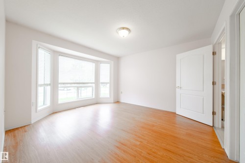 Bright room featuring a large bay window with horizontal blinds, wood-finish flooring, white walls, and a ceiling-mounted light fixture - 120 13320 124 Street Nw, Edmonton, AB 