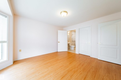 Wood-finish flooring throughout, featuring a large window with blinds, a ceiling-mounted light fixture, and two interior doors, one opening to a tiled bathroom - 120 13320 124 Street Nw, Edmonton, AB 
