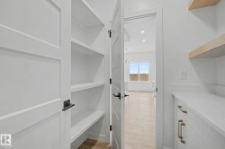 Walk-in pantry featuring built-in shelving, white shaker-style cabinetry, matte black hardware, and light wood-finish flooring - 