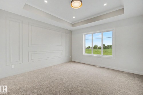 Tray ceiling with recessed lighting and a central flush-mount fixture, large window overlooking a verdant landscape, textured carpet flooring, and decorative wall molding - 2 Willow Heights Drive, Stony Plain, AB 