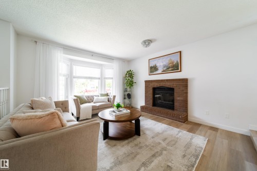 Bright living space featuring a brick fireplace with hearth, wood-finish flooring, and a bay window with ample natural light - 5607 188A Street, Edmonton, AB 