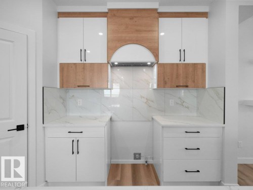 Modern kitchen area featuring a wood-finish range hood, white and wood-finish cabinetry, quartz countertops, a tiled backsplash, and wood-finish flooring - 18723 130 Street, Edmonton, AB 