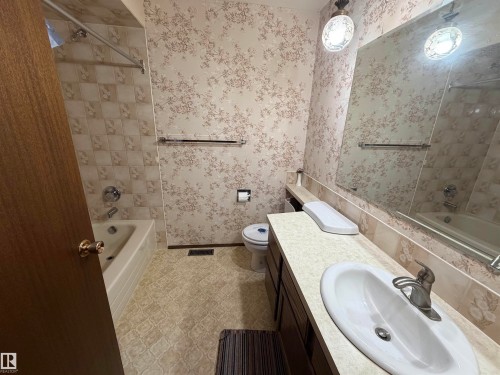 Full bathroom featuring a single vanity with an oval sink, a wall-mounted mirror, a toilet, and a bathtub with a shower fixture and tiled surround - 5319 63 Street, Redwater, AB 