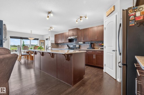 Open-concept kitchen featuring an oversized island with an integrated sink, rich wood-finish cabinetry, light-toned countertops, and dark wood-finish flooring - 74 Santana Crescent, Fort Saskatchewan, AB 