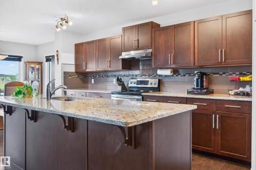 Kitchen featuring a large island with a stainless steel sink and granite countertop, extensive dark wood-finish cabinetry, stainless steel range with overhead vent, and a tiled backsplash - 74 Santana Crescent, Fort Saskatchewan, AB 