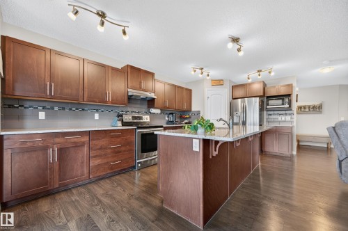Open-concept kitchen featuring wood-finish flooring, rich wood cabinetry, stainless steel appliances, a tiled backsplash, and a large central island with an integrated sink - 74 Santana Crescent, Fort Saskatchewan, AB 