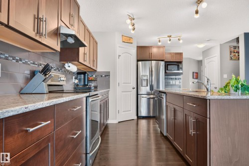 Modern kitchen featuring wood-finish cabinetry, granite-style countertops, stainless steel appliances, a tiled backsplash, and dark wood-finish flooring - 74 Santana Crescent, Fort Saskatchewan, AB 