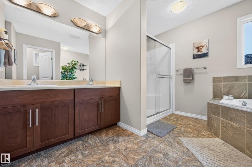 Dual-sink vanity with wood-finish cabinetry and light-toned countertop, featuring a separate shower enclosure with glass doors and a built-in soaking tub with tiled surround, all set against a neutral wall color and patterned floor tiling - 74 Santana Crescent, Fort Saskatchewan, AB 
