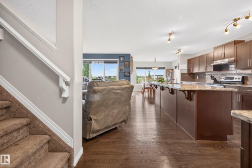 Open-concept living space featuring wood-finish flooring, a kitchen island with seating, dark wood cabinetry, a tile backsplash, and stainless steel appliances - 74 Santana Crescent, Fort Saskatchewan, AB 