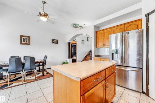 Kitchen island with integrated storage, stainless steel French door refrigerator, wood-finish cabinetry, light-toned tiled flooring, and a ceiling fan with integrated lighting - 6719 164 Ave, Edmonton, AB 