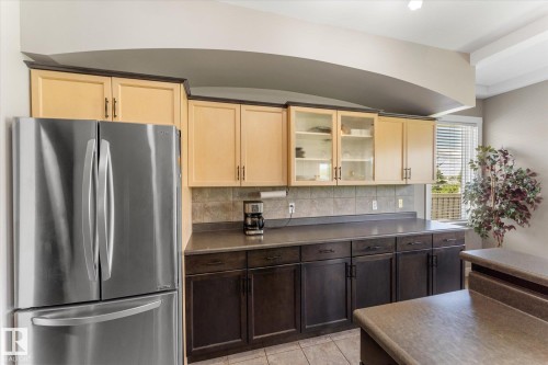 Kitchen featuring two-tone cabinetry with glass-front uppers and dark lower cabinets, stainless steel refrigerator, tile backsplash, and composite countertops - 488 Macewan Road, Edmonton, AB 