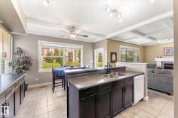 Open-concept kitchen featuring an island with double sink, dark wood-finish cabinetry, tile flooring, and a dining area with a large window - 